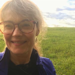 Woman's head, with red glasses, blond hair, smiling, green field behind her